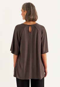 Bruine top met korte mouwen en een keyhole-detail aan de achterkant. Soepele stof, relaxte pasvorm en gebogen zoom. Gecombineerd met losse, donkere broeken.