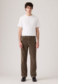 T-shirt bianco a maniche corte abbinato a pantaloni in velluto a coste marroni. I pantaloni presentano una consistenza visibile e una vestibilità a gamba dritta, indossati con scarpe nere.