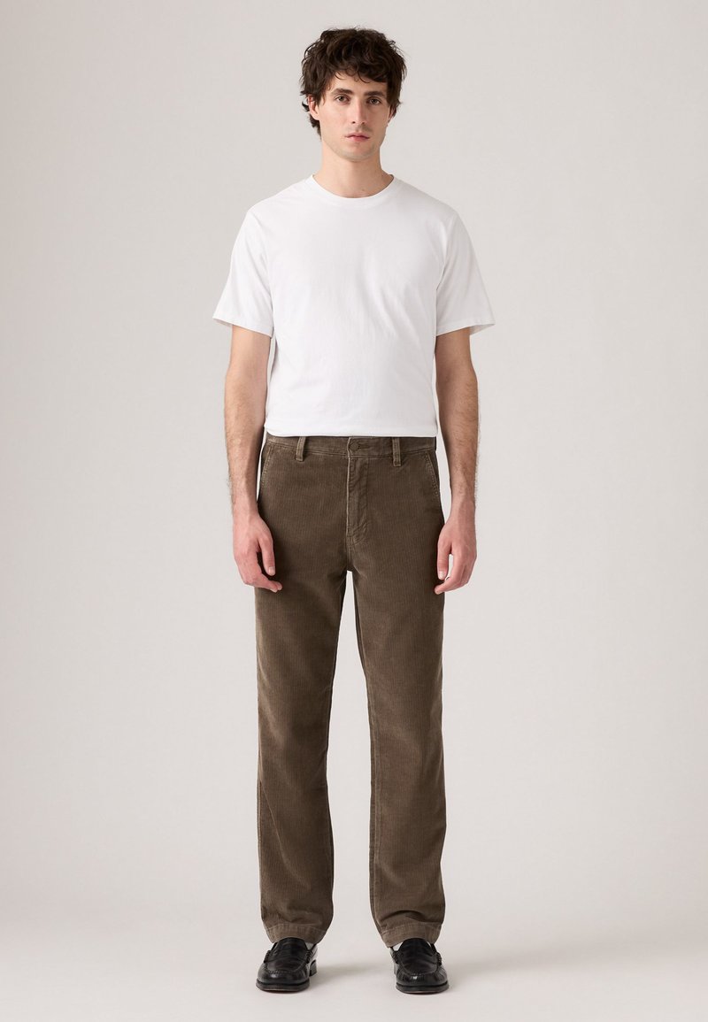Weißes Kurzarm-T-Shirt kombiniert mit braunen Cordhosen. Die Hosen weisen eine sichtbare Textur auf und haben einen geraden Schnitt, getragen mit schwarzen Schuhen.