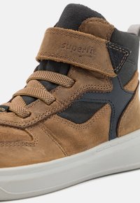 Zapato deportivo alto de ante marrón y tela con acentos en negro, sobrecapas texturizadas y una correa de Velcro, con una suela de goma blanca y cordones.
