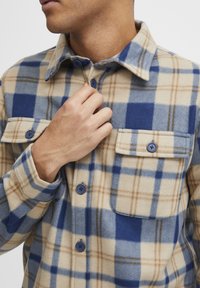 Fleece shirt in een geruite patroon met blauwe, beige en bruine tinten. Ontworpen met een shirtkraag, voorknopen en twee borstzakken.