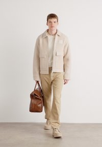 Giacca di pile beige con due tasche frontali, camicia crema sotto, pantaloni khaki e scarpe color nocciola. Borsa in pelle marrone in mano.