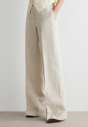 Personne portant un pantalon beige à jambes larges et des chaussures marron pointues, main dans la poche, debout contre un mur uni.