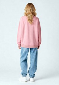 Felpa oversize rosa chiaro con spalle scese, polsini e orlo a coste, abbinata a jeans azzurri larghi e sneakers bianche.