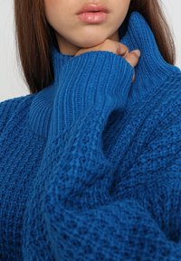Pull en tricot bleu avec un col montant côtelé et un motif texturé. Le tissu semble doux, avec des fils lâches créant une apparence confortable.