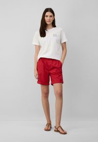 T-shirt blanche en coton avec texte sur la poitrine, associée à un short rouge avec une taille élastique et des ourlets retroussés. Des sandales marron complètent la tenue.