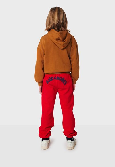 Felpa marrone tagliata con cappuccio, abbinata a pantaloni della tuta rossi con lettere blu. Sneakers bianche con dettagli neri completano il look.