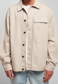 Mężczyzna w beżowej kurtce zapinanej na guziki z klapką na piersi i dwoma dolnymi kieszeniami, w połączeniu z jasnoniebieskimi dżinsami i białą koszulą pod spodem.