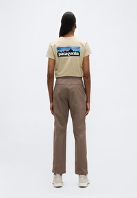 Άτομο που στέκεται με την πλάτη προς τα  εμπρός φορώντας μπεζ μπλουζάκι Patagonia, καφέ παντελόνι και άσπρα αθλητικά παπούτσια σε απλό φόντο.