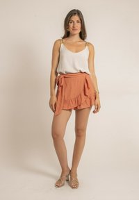 Haut blanc à bretelles spaghetti et accessoires en or décoratifs, associé à une jupe portefeuille orange avec un ourlet à volants. Sandales à talons argentées.