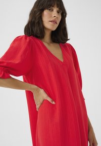 Robe rouge texturée avec un décolleté en V et des manches bouffantes. Poche latérale visible, mettant en valeur un tissu léger et une coupe décontractée.