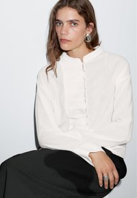 Witte blouse met een hoge kraag, voorzien van knoopdetails en subtiele plooien. Model draagt een zwarte rok en een zilveren ring, met een minimalistische achtergrond.