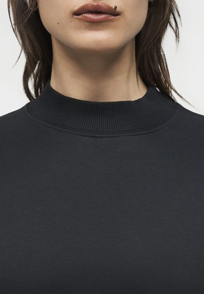 Κοντινό πλάνο ενός ατόμου που φοράει μαύρο πουκάμισο με ψηλό γιακά, με εμφανή τα χείλη, το πηγούνι, τον λαιμό και μια φακίδα στο πρόσωπο πάνω από τα χείλη.