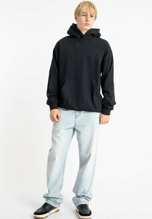 Czarna bluza z kapturem z przednią kieszenią, noszona z jasnoniebieskimi dżinsami i czarnymi trampkami z białą podeszwą. Gładka tekstura tkaniny.