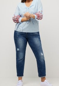 Blouse rayée bleu clair avec des manches bouffantes et broderies florales, détail nœud ; associée à un jean foncé déchiré et des baskets blanches.