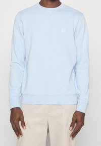 Ljusblå bomullströja med rundad halsringning, ribbade ärmslut och en fyrkantig logotyp på vänster bröst, med en avslappnad passform.