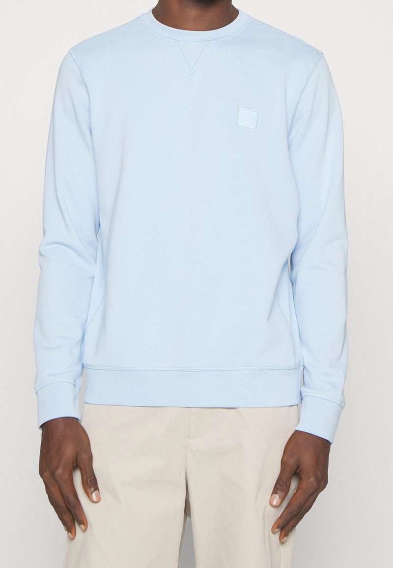 Ljusblå bomullströja med rundad halsringning, ribbade ärmslut och en fyrkantig logotyp på vänster bröst, med en avslappnad passform.