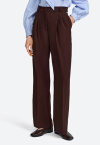 Bruine geplooide broek met hoge taille, zachte stof, wijd uitlopende pijpen en zijzakken, gecombineerd met zwarte platte schoenen.