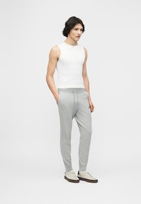 Νεαρός άντρας που στέκεται φορώντας λευκό αμάνικο μπλουζάκι, ανοιχτό γκρι φόρμα και μπεζ αθλητικά παπούτσια σε απλό ανοιχτό γκρι φόντο.