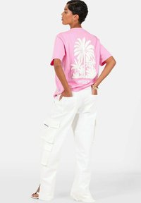 Roze katoenen T-shirt met een witte palmboomafbeelding op de achterkant, gecombineerd met witte cargobroeken met meerdere zakken en zijsplitsingen.