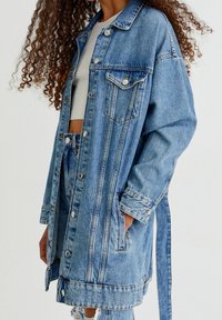 Veste en denim de couleur bleu clair, coupe oversize, fermeture par boutons, deux poches poitrine, et un détail de lien à la taille. Présente une légère décoloration.