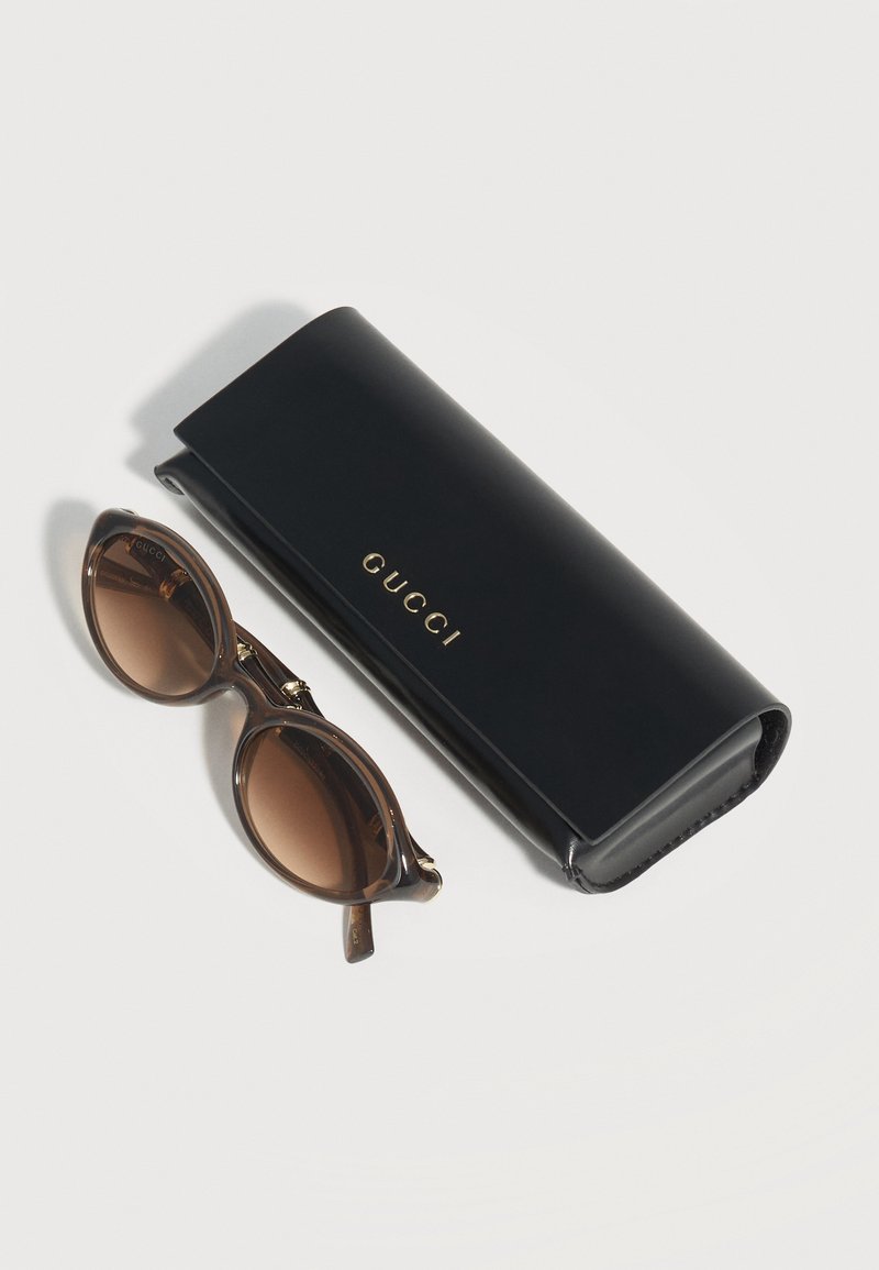 Occhiali da sole ovali marroni con lenti sfumate accanto a una custodia Gucci nera con logo dorato su una superficie bianca.