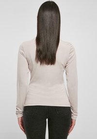 Langsleeve lichtbeige top gemaakt van zachte stof, aansluitend ontwerp met een rechte zoom. Gecombineerd met donkergrijze aansluitende jeans, gezien van achteren.