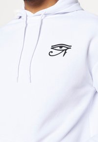 Persona che indossa una felpa con cappuccio bianca con un simbolo nero dell'Occhio di Horus sul petto sinistro, primo piano di parte superiore del torso e collo.