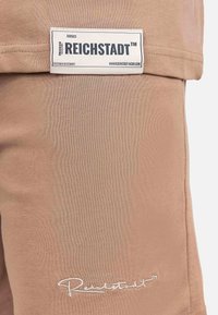 Etichetta in tessuto beige con marchio "REICHSTADT", caratterizzata da testo bianco ricamato e texture liscia. Cuciture di accentuazione visibili vicino ai bordi.