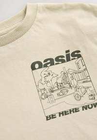 Camiseta de algodón color beige con cuello redondo, que presenta un dibujo en línea negra de una escena con árboles, personas y un scooter, junto con el texto "oasis ESTAR AQUÍ AHORA."