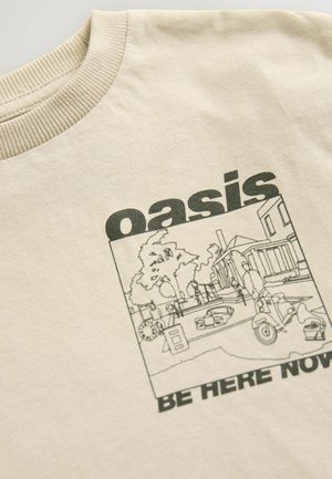 Väri vaalean beige puuvillapaita, jossa on pyöreä kaula-aukko. Paidassa on mustaa viivataidetta, jossa näkyy maisema puita, ihmisiä ja skootterin, sekä teksti "oasis BE HERE NOW."