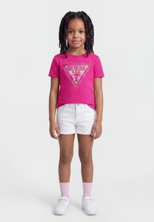 Jeune fille aux cheveux tressés portant un t-shirt rose GUESS, un short blanc, des chaussettes roses et des baskets blanches, debout devant un fond uni.
