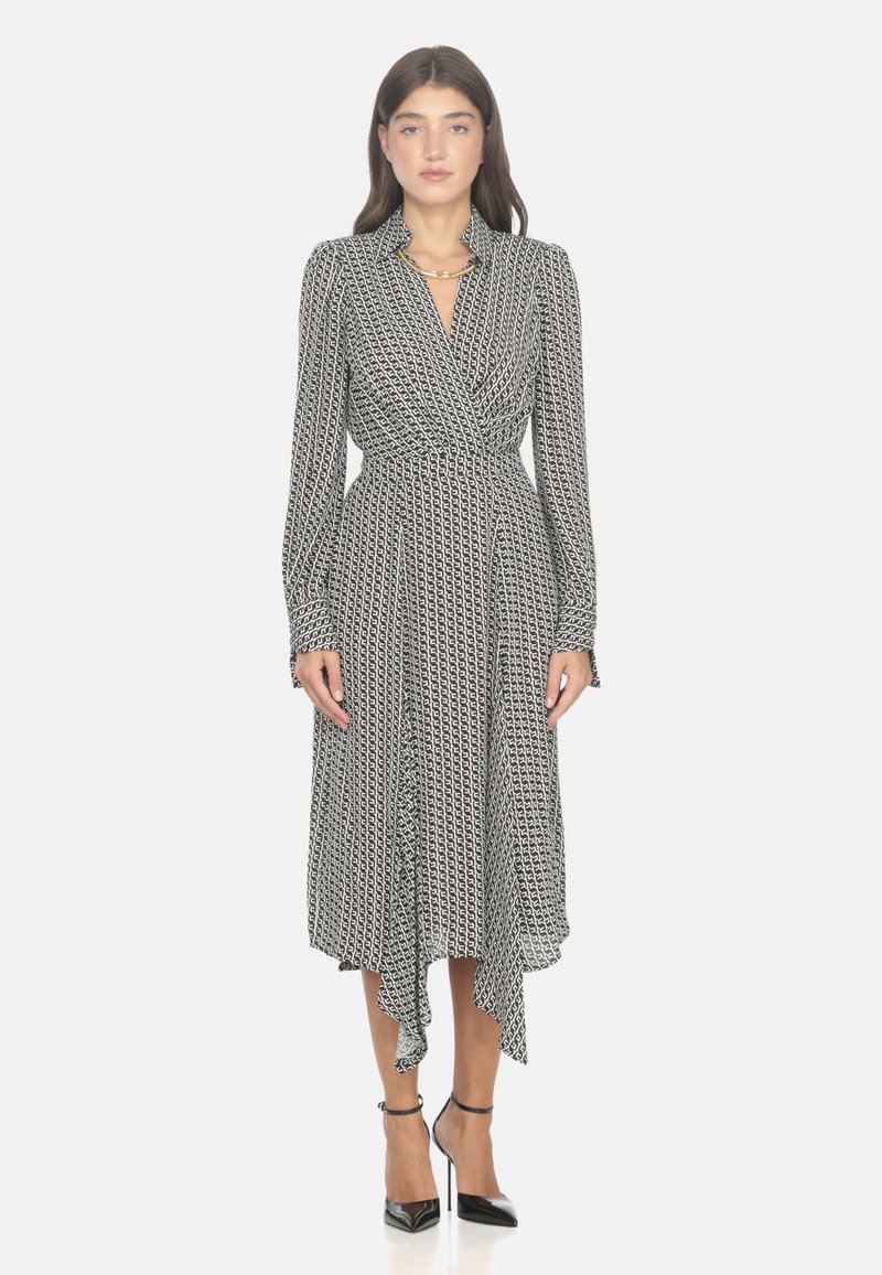 Vestito avvolgente a righe in bianco e nero con maniche lunghe e orlo arricciato, caratterizzato da una vita aderente e dettagli con bottoni. Indossato con tacchi neri.