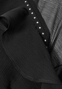 Top in tessuto nero con frange testurizzate e una linea decorativa di piccole perle bianche lungo un lato. La lucentezza liscia contrasta con la texture.