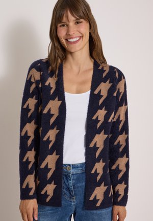 Femme souriante portant un cardigan bleu marine et beige à motifs par-dessus un haut blanc et un jean bleu sur un fond neutre.