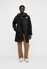 Μαύρο αδιάβροχο μπουφάν με κουκούλα, με δύο μπροστινές τσέπες και φερμουάρ κλεισίματος. Φορεμένο πάνω από μαύρο πουκάμισο και μπεζ παντελόνι.