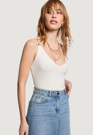 Jeune femme aux cheveux châtain clair portant un haut blanc sans manches à bretelles chaîne, des colliers en or superposés et une jupe en jean bleue taille haute.