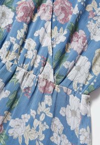 Tissu bleu à motif floral avec de grandes fleurs blanches et roses et des feuilles vertes, plissé au niveau d'une couture au centre.