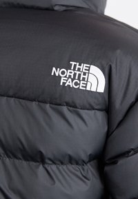 Schwarze Daunenjacke aus strukturiertem Material, mit gesteppten Bereichen und einem weißen, gestickten Logo von The North Face auf der oberen linken Seite.