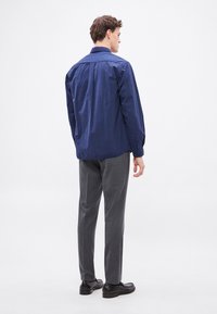 Cămașă albastru marin cu mâneci lungi, material ușor. Pantaloni gri cu model, croială ajustată, asortați cu pantofi negri. Vedere din spate.