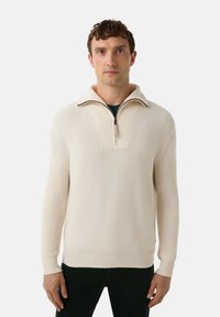 Mann trägt einen cremefarbenen, gerippten Pullover mit Viertel-Reißverschluss und Kragen, steht vor einem schlichten weißen Hintergrund.