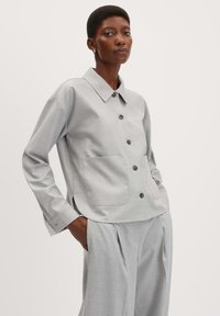 Veste gris clair à boutons avec col. Présente deux poches avant et des manches longues avec poignets. Texture lisse, léger motif visible.
