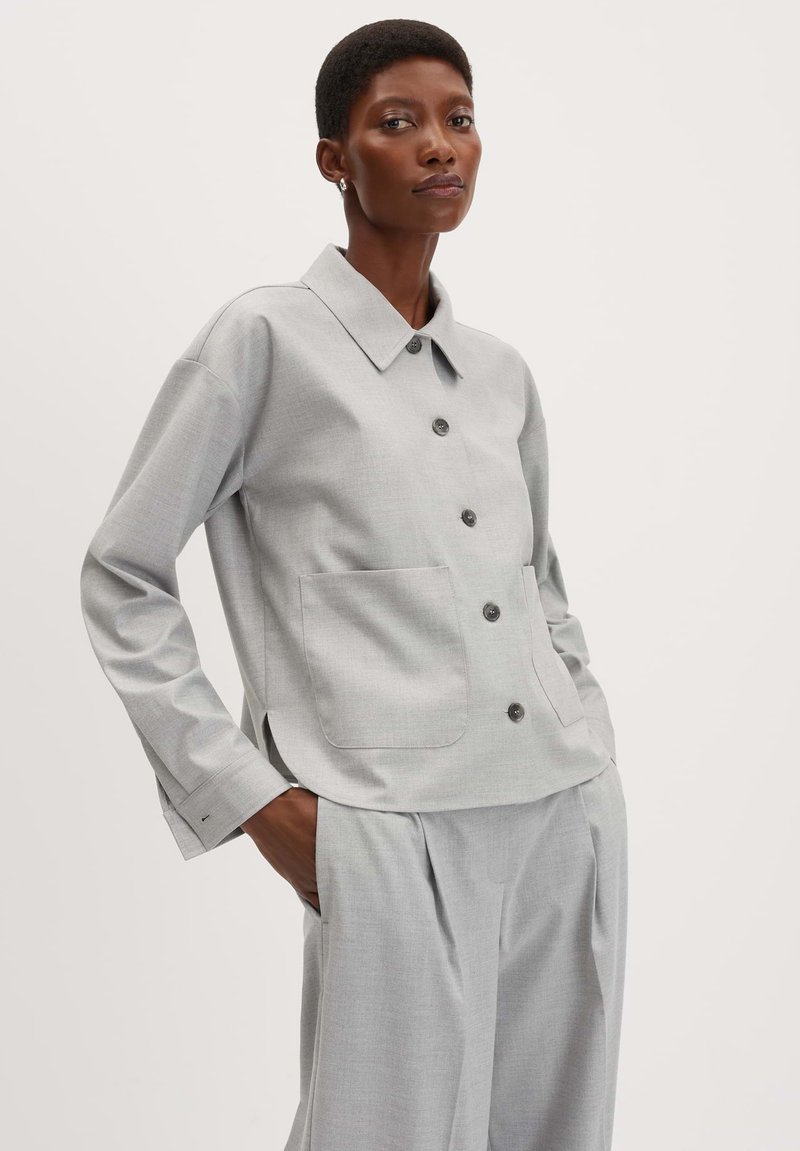 Veste gris clair à boutons avec col. Présente deux poches avant et des manches longues avec poignets. Texture lisse, léger motif visible.
