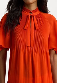 Red fabric top with pleated texture, high neckline, and bow tie detail. Short sleeves with ruffled edges. Smooth, lightweight material.