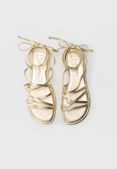 Gylne metalliske flate sandaler med flere tynne stropper og en rund ankelbånd. Glatt tekstur med firkantet tåform og blomsterdesign på innersålen.