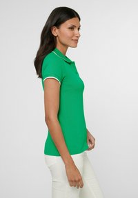 Groene polo met witte bies op de kraag en mouwen, gemaakt van een gladde stof, met een getailleerd ontwerp, gefotografeerd in zijprofiel tegen een effen achtergrond.