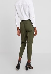 Pantalons fuselés vert olive avec une ceinture extensible, dotés de poches latérales. Associés à une chemise blanche à manches longues et des talons ouverts noirs.