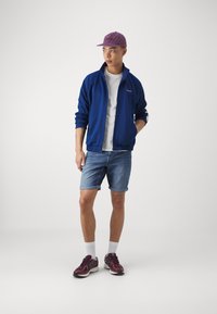 Plava jakna na zip, bijela majica, skraćene traperice, ljubičasta kapa i bordo sportske cipele s bijelim čarapama. Opušteni outfit.