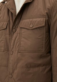 Giacca isolante marrone con chiusura a bottoni, due tasche sul petto con patta e una texture liscia e leggermente lucida.