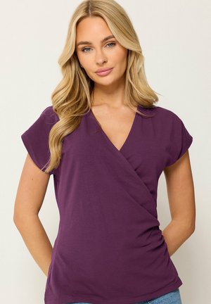 Femme aux longs cheveux blonds portant un haut violet à manches courtes de style cache-cœur et un jean bleu sur un fond uni.